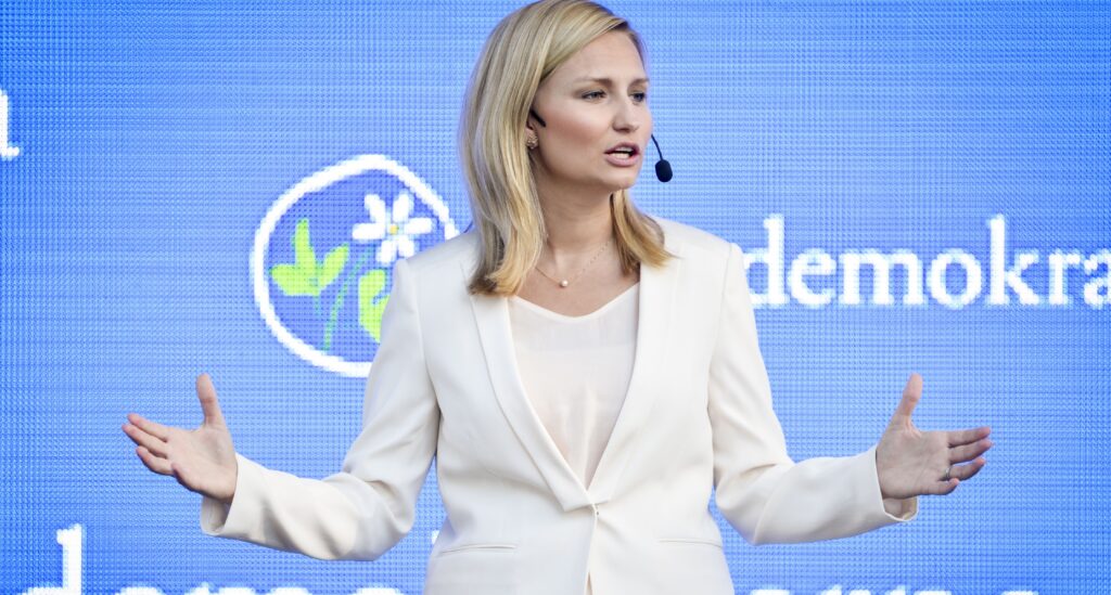 Kristdemokraternas ledare Ebba Busch Thor har vita kläder och sträcker ut armarna framför en ljusblå bakgrund där det står Kristdemokraterna.