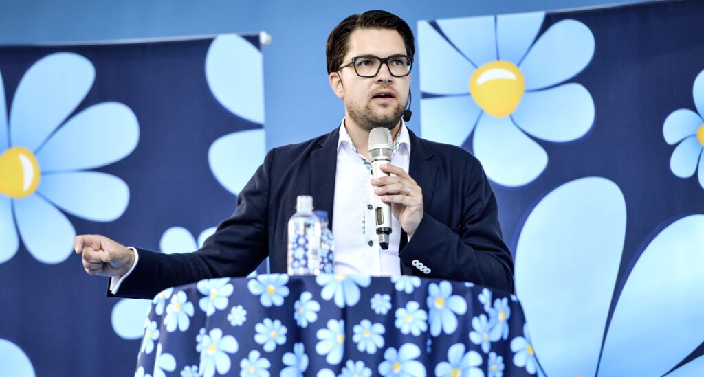 Jimmie Åkesson står bakom ett bord och håller i en mikrofon som han pratar i. På bordet ligger en duk med blåsippor och i bakgrunden finns ett draperi med blåsippor. Blåsippor är Sverigedemokraternas symbol för partiet.