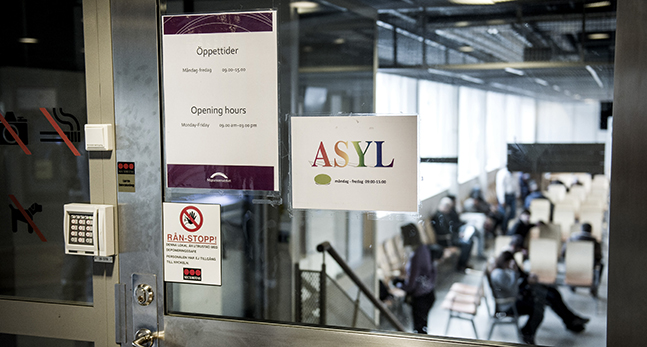En fönsterdörr. På fönstret finns lappar där det står Asyl och öppettider. Det finns också en liten lapp med en överkorsad hand på.
