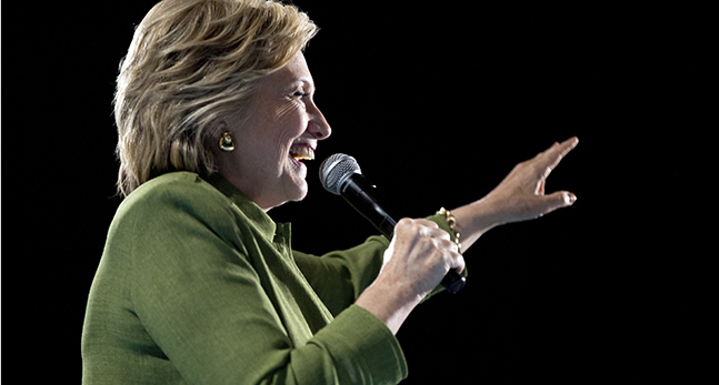 Hillary Clinton i halvfigur och i profil. Hon håller en mikrofon och ser glad ut.