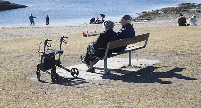 Två äldre personer sitter på en bänk vid en sandstrand och pratar.