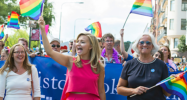 Ebba Busch Thor i en cerise topp viftar med en flagga med regnbågens färger åt människor när hon går i paraden.