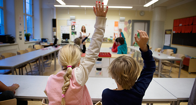Ett klassrum med barn som räcker upp handen. Man ser barnens ryggar. Längst fram ser man en lärare stå vid whiteboarden.