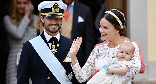 Prins Carl Philip och prinsessan Sofia lämnar kyrkan med sin son Alexander. Carl Philip har uniform. Sofia en vit spetsklänning och hårband av pärlor. Alexander i hennes famn har en vit dopklänning. Sofia vinkar.