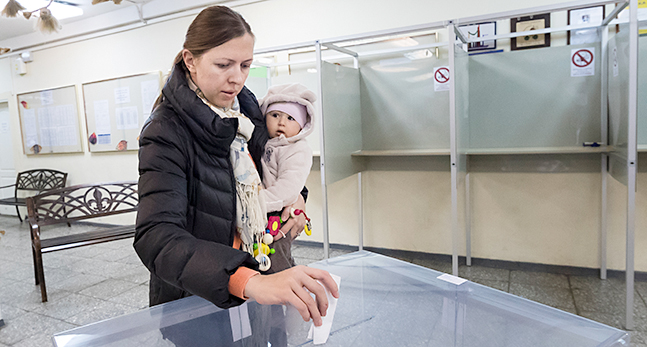 En kvinna med en bebis på armen lägger en lapp i en låda av genomskinlig plast.