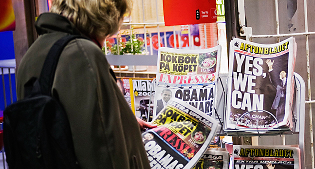 en kvinna läser en kvällstidning. Hon står framför ett stånd med flera olika tidningar.