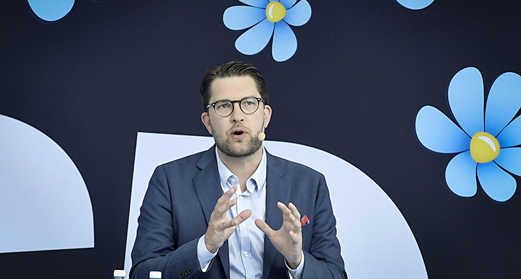 Jimmie Åkesson står bakom en vägg med partiets symbol, blå blommor.