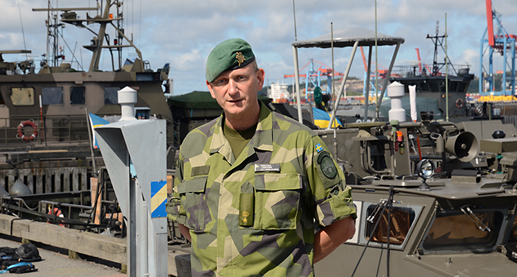 Thomas Olsson står i kamouflagekläder och grön basker. Han här händerna bakom ryggen. Bakom honom ser man en båt.