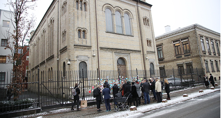 En bild på byggnaden som är synagogan i Göteborg. Utanför staketet står en samling människor och tittar på staketet där det hänger pappershjärtan.