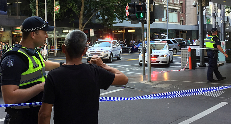 En bild på en gata i Melbourne. Längst fram i bilden syns en polis som pratar med en förbipasserande. Ett band spärrar av gatan mellan dem. Längre bak i bilden syns polisbilar med sirenerna påslagna.