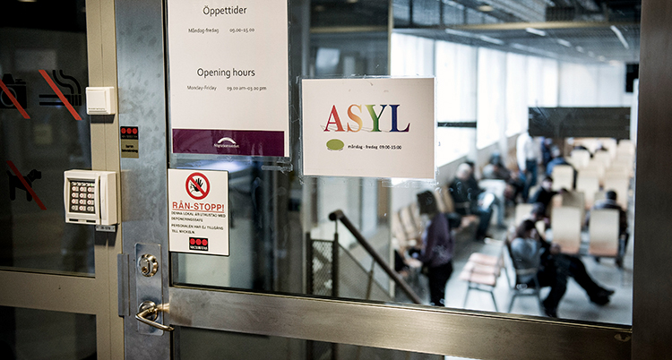 Skylt på en glasdörr där det står Asyl. Innanför väntar människor sittande på stolar.