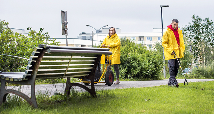 Två ungdomar plockar skräp med långa gripklor vid en gräsmatta. Det regnar och ungdomarna har på gula regnjackor.