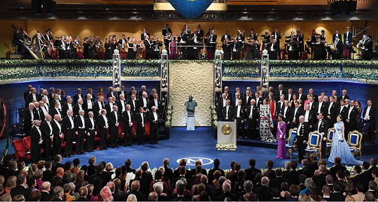 En bild från Konserthuset och förra årets prisutdelning. Vi ser hela scenen ovanifrån, nobelpristagarna i stolar till vänster, kungafamiljen till höger. i mitten står en byst av alfred nobel.