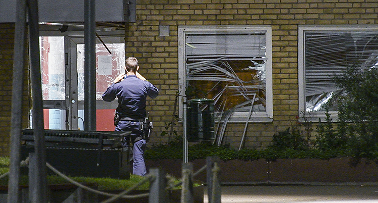En polis står framför en port till ett tegelhus. Fönstret till entredörren är krossad. Likaså fönstret bredvid dörren.