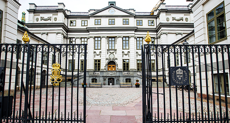 Fasaden av ett gammalt imponerande stenhus i vitt med fyra pelare i entren. Framför huset en stenlagt gård som inramas av ett svart högt staket i järnsmide med guldknoppar.