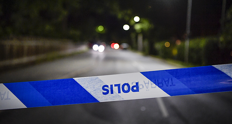 Ett blåvitt plastband med ordet polis i förgrunden av bilden. Bakgrunden visar en tom asfalterad väg i mörker.
