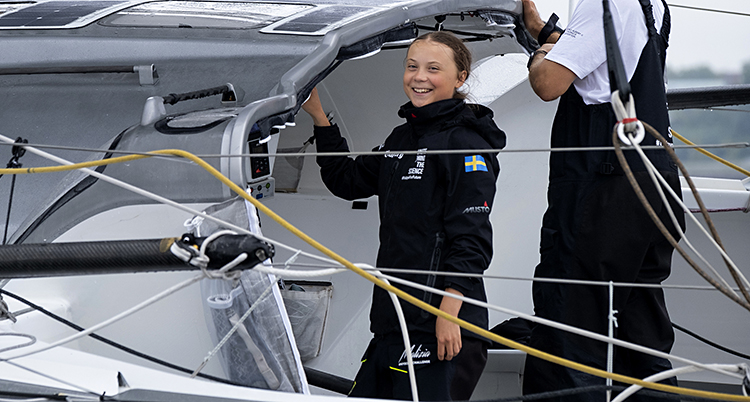Greta Thunberg i mörkblå vindkläder står på segelbåten och skrattar.
