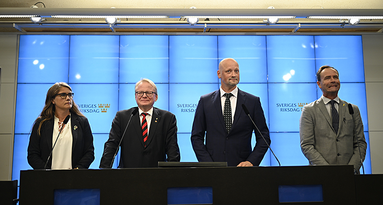 Fyra politiker står i ett rum i riksdagen och pratar med journalister. Längst till vänster står en kvinna. Hon har blus och kavaj. De andra tre är män. De har kostym och slips. De har varsin mikrofon som de kan prata i.