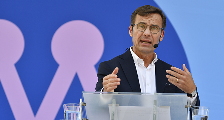 Kristersson håller händerna framför sig samtidigt som han talar. Han är i en talarstol, ser allvarlig ut. Moderaternas märke är på väggen.