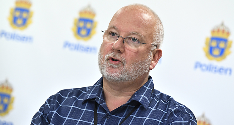 En äldre man med glasögon och vitt skägg framför en vägg med polisernas symbol.