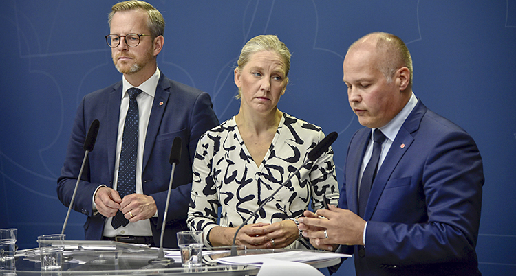 De tre ministrarna står på rad famför ett bord. Morgan Johansson talar.