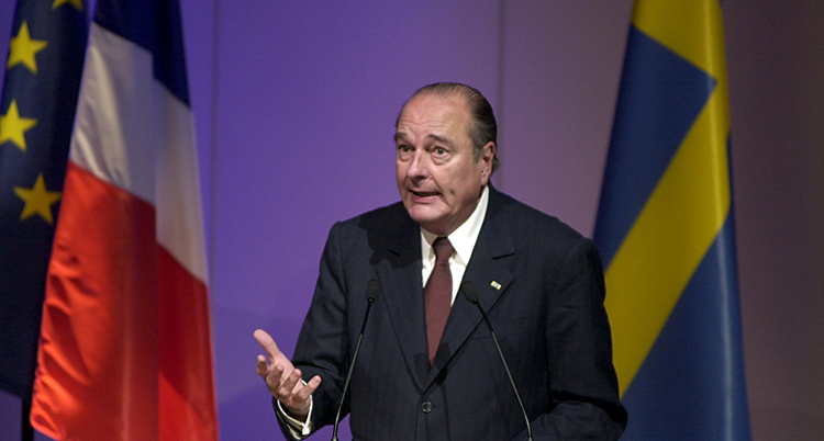 Bilden är från år 2000. Den dåvarande franske presidenten Jacques Chirac står på en scen och pratar. Han har kostym och slips. I bakgrunden syns den franska flaggan och den svenska flaggan.