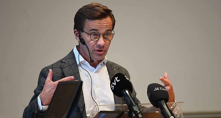 Ulf Kristersson håller ett tal. Han står och pratar i flera mikrofoner. Han gör gester med händerna medan han pratar. Han har glasögon, vit skjorta och en kavaj.