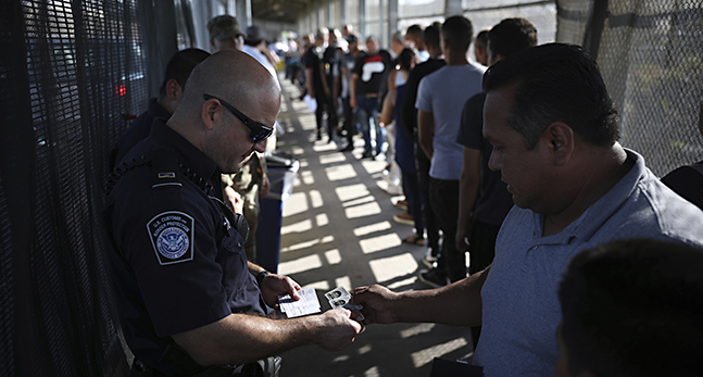 En polis tittar på en mans id-kort. Framför mannen ringlar kön lång. De omges av höga stängsel.