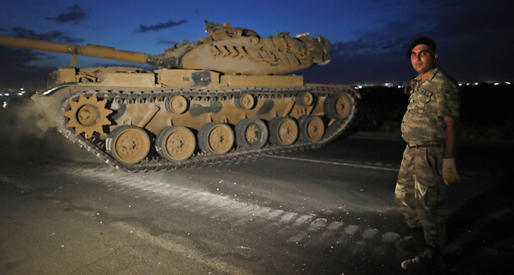 Bilden är tagen på kvällen eller på natten. Det är mörkt. Vi ser en bandvagn. En militär står bredvid bandvagnen. Han har en basker och kläder i kamouflage-färger.