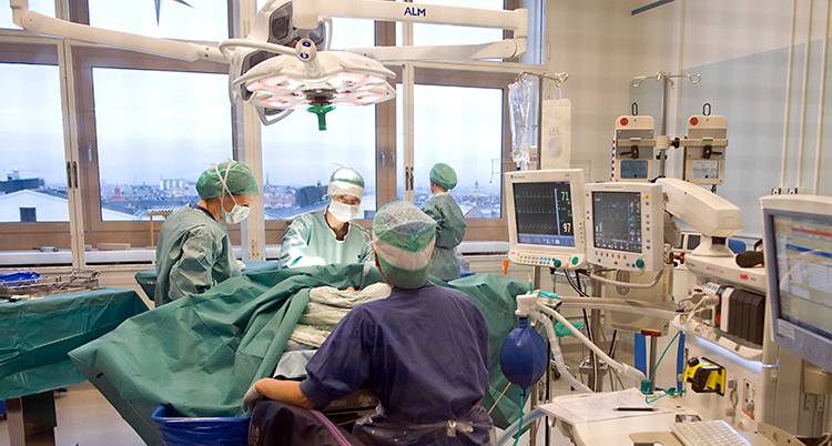 Bilden visar en operationssal på ett sjukhus. Flera personer jobbar med en patient. De som jobbar har på sig skyddskläder och skydd för munnen. Runt om finns en massa instrument och skärmar.