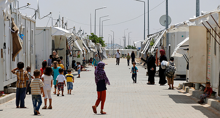 Bilden visar ett läger för flyktingar i Turkiet. Där finns flyktingar från Syrien. På en gata syns barn och kvinnor. Längs gatan finns små byggnader.