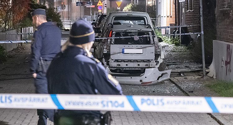 Poliser har spärrat av en gata med blåvita band. En polisrygg i förgrunden. En skadad bil i bakgrunden.