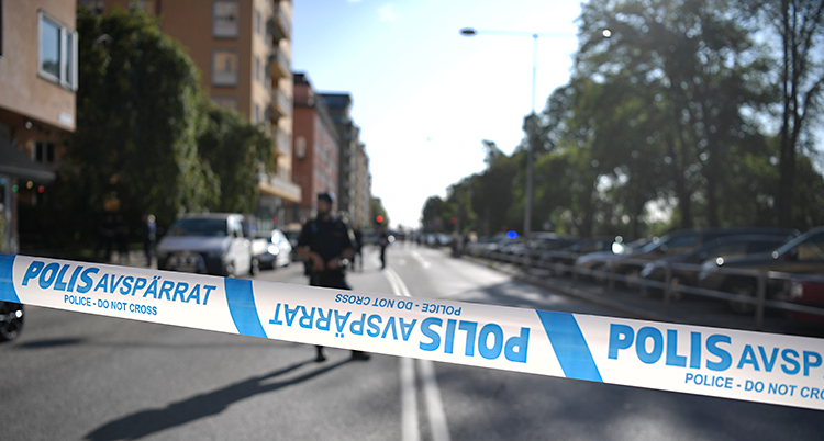 Bilden visar en gata i Stockholm. På ena sidan av gatan syns en rad med lägenhetshus. Polisen har satt upp ett avspärrningsband. I bakgrunden syns en polis som vaktar.