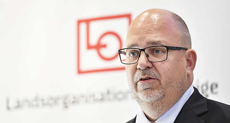 En man i 50-årsåldern som står framför en vägg där det står Landsorganisationen i Sverige. Mannen är flintskallig med kortklippt hår på sidorna. Han har glasögon och ett kort skägg runt munnen.
