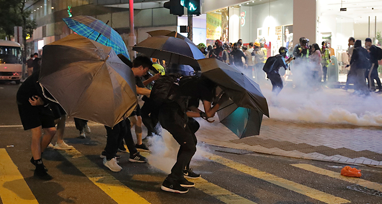 En gata i Hongkong på kvällen. Det syns tårgas på gatan. Längre bort syns poliser. Närmaste i bild står några människor med paraplyer.