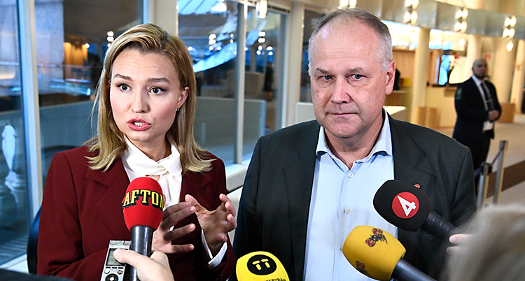 Ebba Busch Thor och Jonas Sjöstedt blir intervjuade tillsammans av flera journalister. Journalisterna sträcker fram mikrofoner mot dem. Ebba Busch Thor har långt blont hår och en röd kavaj. Jonas Sjöstedt har kort grått hår och en svart kavaj.