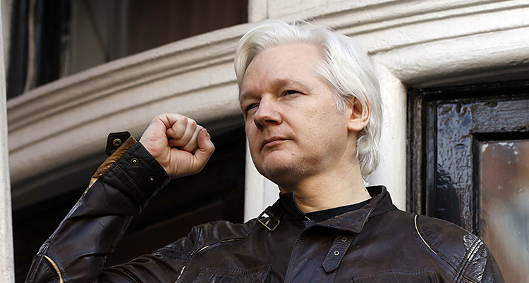 Julian Assange håller upp en näve i luften. Han har grått hår och en jacka av skinn på sig.