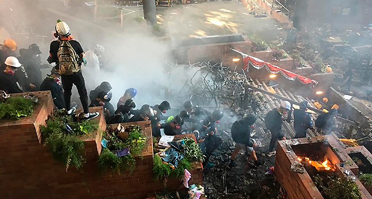 En bild från trappan utanför ett universitet i Hongkong. Flera personer går nerför trappan. Flera har hjälmar på sig. Det är rök i luften.