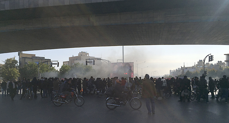 En stor samling med människor i staden Isfahan. Framför folksamlingen kör två personer förbi med mopeder. Bakom folksamlingen syns rök i luften.