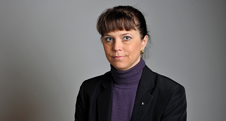En kvinna tittar in i kameran. Hon har mörkt hår som är uppsatt. Hon har lugg. Hon har en lila polo och en mörk kavaj. Bilden är tagen i ett rum med grå vägg i bakgrunden.