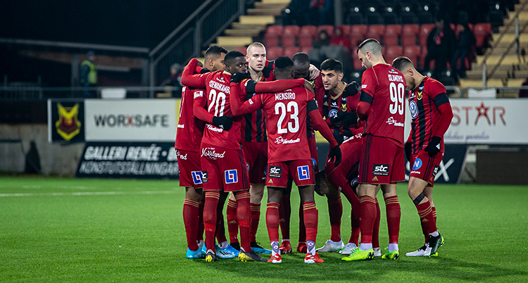 Bilden visar en fotbollsplan. På planen står en grupp spelare i ring och pratar med varandra. De har röda tröjor och röda shorts.