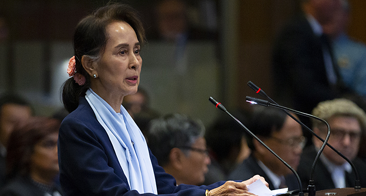 Aung San Suu Kyi står i en sal i en domstol och pratar i en mikrofon. Hon har ett hårband i nacken och en ljusblå sjal.