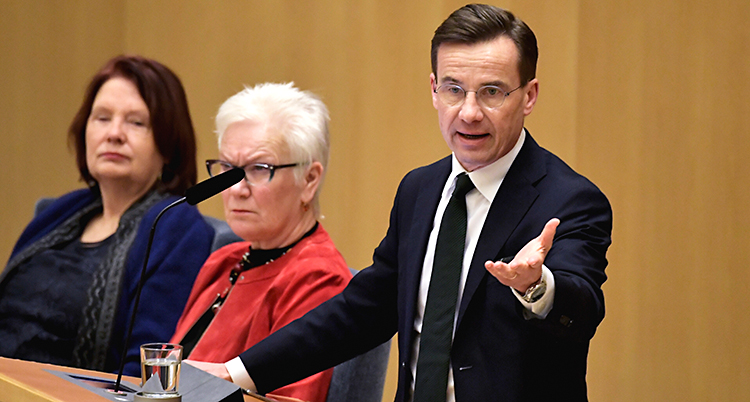 Ulf Kristersson pratar i en mikrofon. Han står i en talarstol i Sveriges riksdag. Han har glasögon och kostym.
