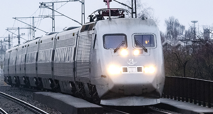 Ett tåg från SJ rullar på en räls. Det är ett grått tåg som heter X2000. Vädret är också grått.