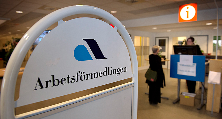 Bilden är från en Arbetsförmedling. Längst fram i bilden finns en skylt där det står Arbetsförmedlingen. Längre bort står en person som får hjälp av en arbetsförmedlare.