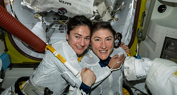 Jessica Meir och Christina Koch håller om varandra i stationen i rymden. De har vita dräkter på sig.