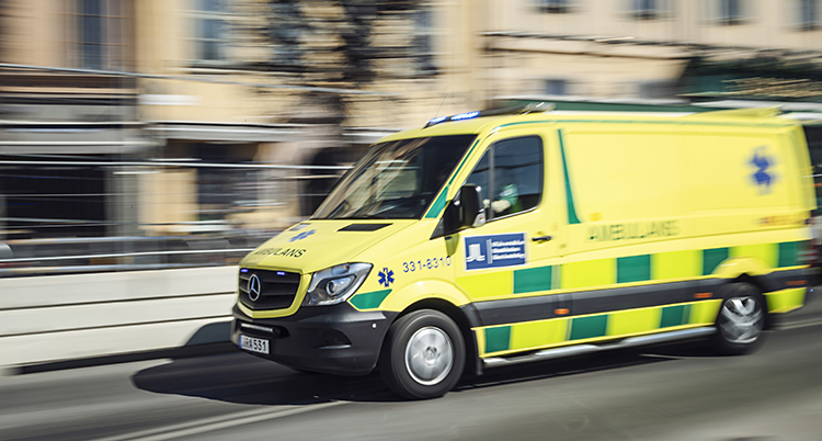 En ambulans som åker på en gata. Bilden är lite suddig för att visa att ambulansen åker fort. Ambulansen är gul med gröna detaljer.