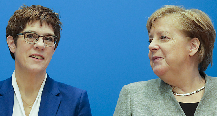 De två tyska politikerna Annegret Kramp-Karrenbauer och Angela Merkel står bredvid varandra. Annegret står till vänster. Hon har glasögon och en blå kavaj. Hon tittar rakt fram. Angela Merkel står till höger. Hon har en grön kavaj. Hon tittar på Annegret.