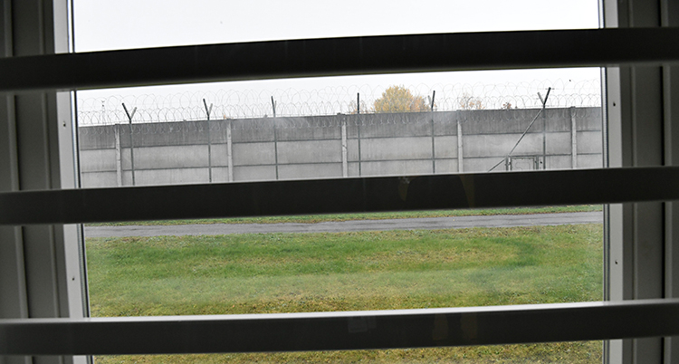 Bilden är tagen från en cell på ett fängelse. Ut genom fönstret. Det finns ribbor för fönstret. Längre bort syns en mur med taggtråd.