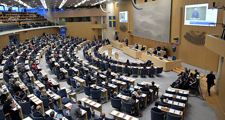 En bild som visar den stora salen i riksdagen. Politiker sitter i sina bänkar och lyssnar. En politiker står längst fram och pratar.